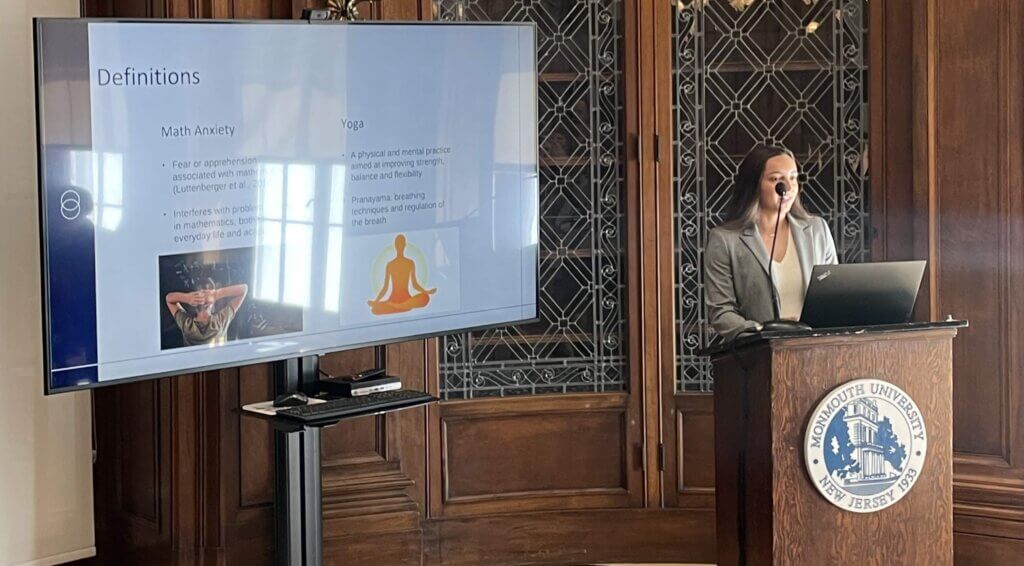 Student give a presentation from a podium, with slides shown on a monitor to her side