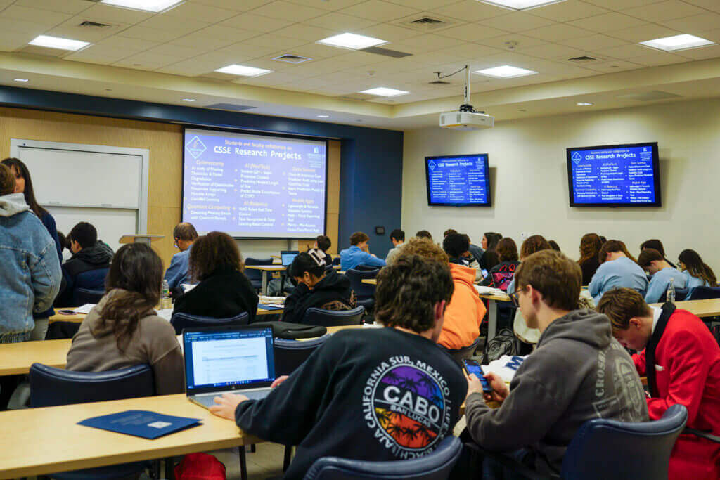 Students working at tables in a classroom. Monitors on the wall show a slide labeled CSSE Research Projects