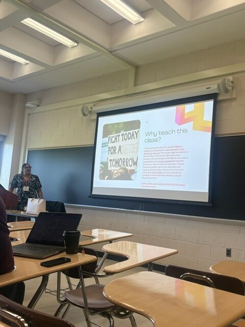 Woman presenting slides in a classroom. Slide titled "Why teach this class?", photo in slide showing protester holding sign that reads "Fight Today For a Better Tomorrow"