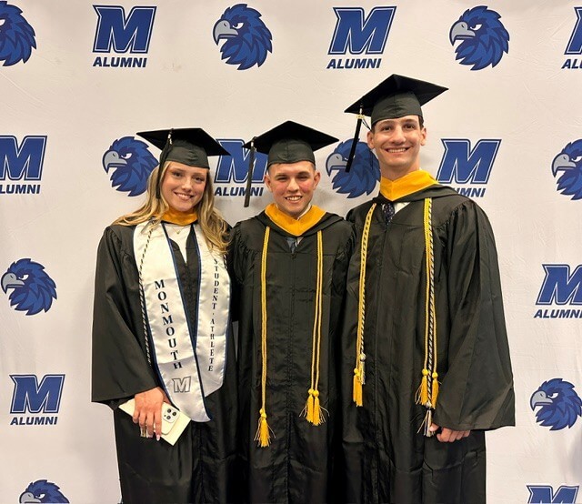 Three graduates in caps and gowns standing in front of banner with Monmouth University insignia