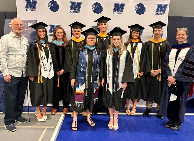 Group of graduates in caps and gowns alongside faculty, posing for photo in front of Monmouth banner.