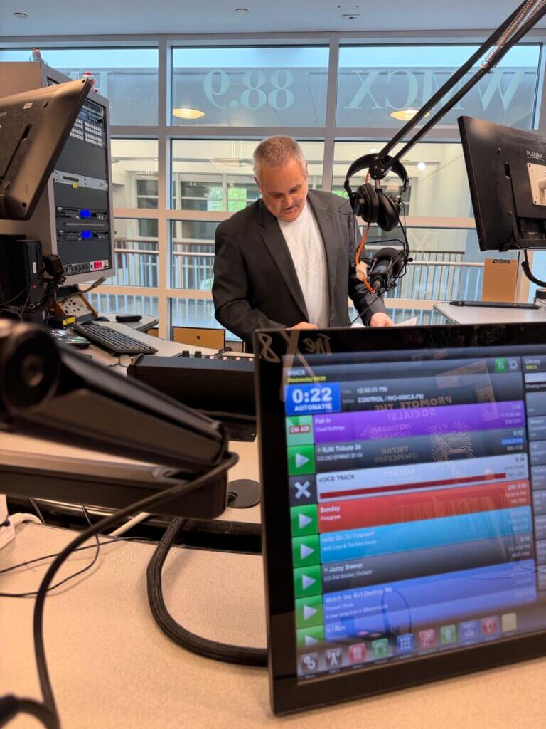 Man at radio jocky desk, microphone on an arm near him. Monitors and audio equipment