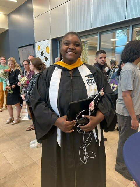 Student in graduate gown, holding hat and degree in hand.