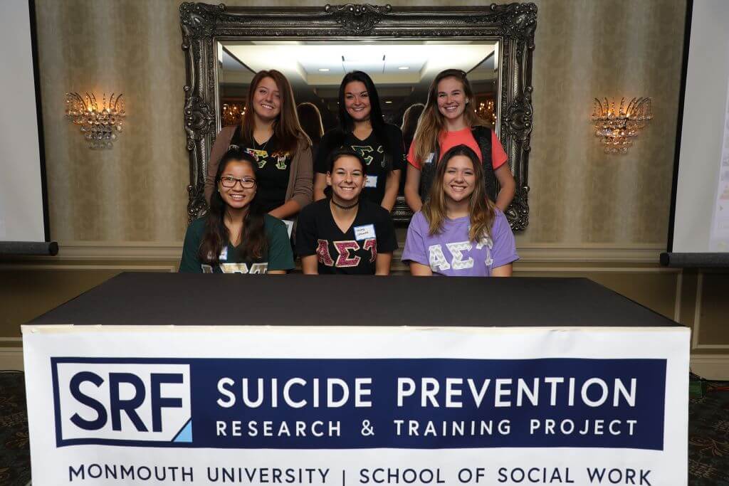 Six students are behind a table with a banner on it that reads SRF, Suicide Prevention Research & Training Project. Monmouth University School of Social Work. 