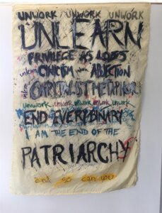 Painted scroll featuring text in various colors, strokes, and sizes. Roughly, it reads as follows: "Unwork Unwork Unwork. Unlearn privilege as loss, unlearn cynicism, unlearn abjection, unlearn capitalist metaphor, unwork unwork unwork unwork unwork. End every binary. I am the end of the patriarchy and so can you." 