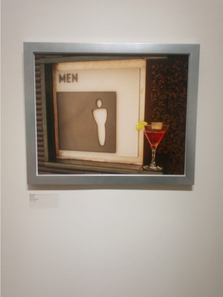 Framed color photo of a men's bathroom sign with a cocktail drink positioned to it's right.