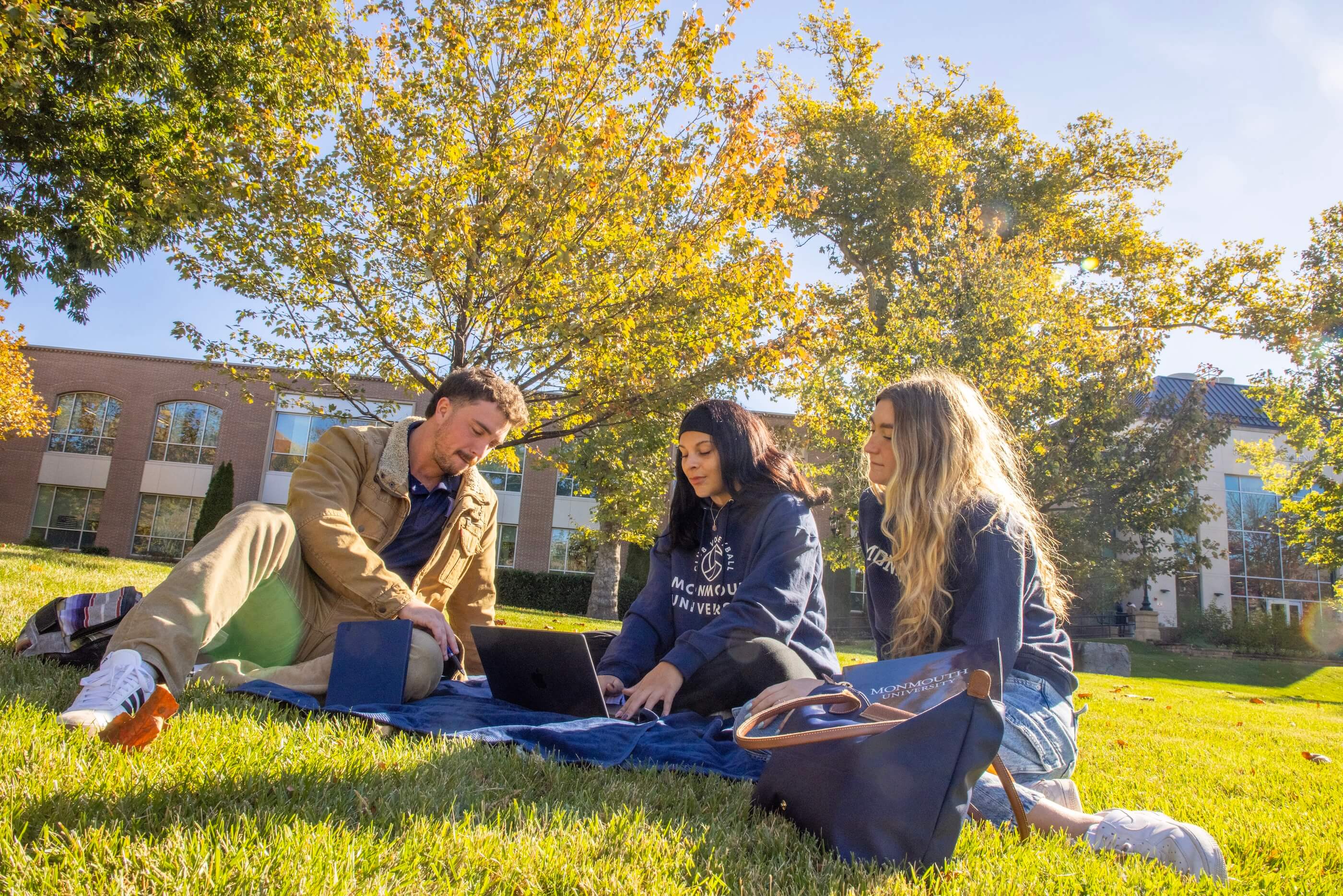 Three students sitting on a campus lawn, trees with fall leaves and a school building behind them