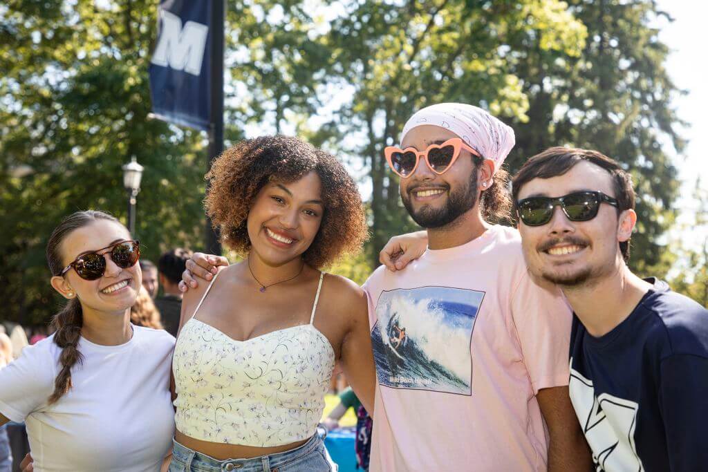 Four students standing together and smiling at the Involvement Fair on a sunny day at Shadow Lawn.