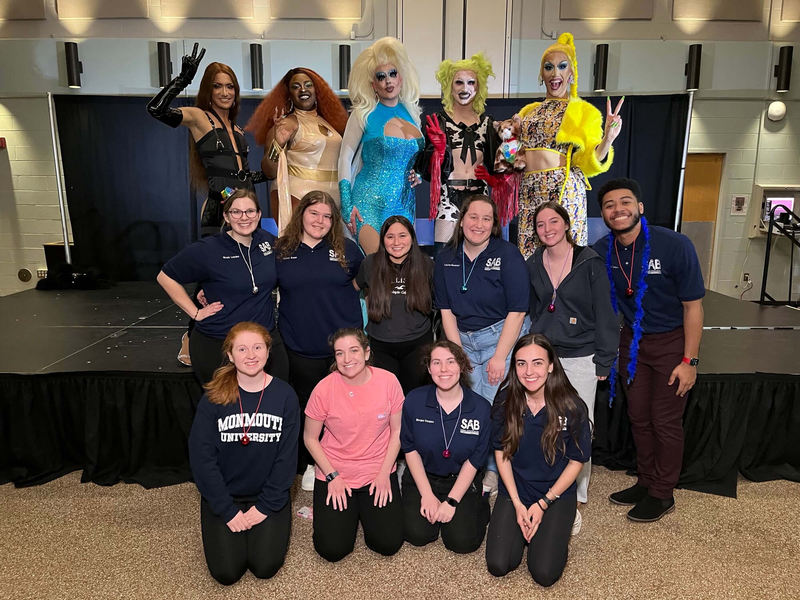 A group of people wearing SAB branded shirts posing in a group with people in drag.