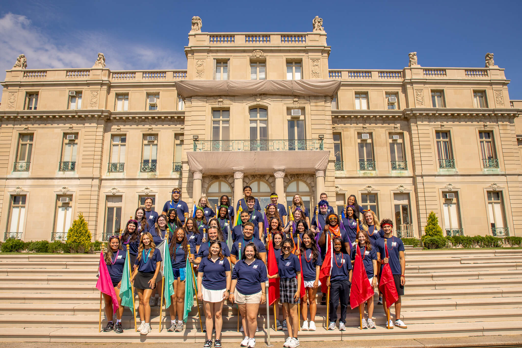 Young adults in blue polos bearing the Monmouth University logo, holding red, yellow, green, orange, purple and burgundy flags