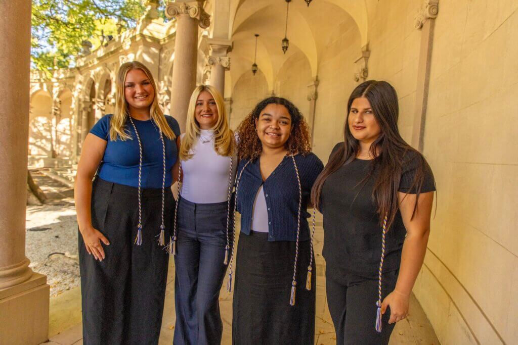 Four young women wearing gold and blue tassels over their shoulders, posing by stone columns in an outdoor garden.