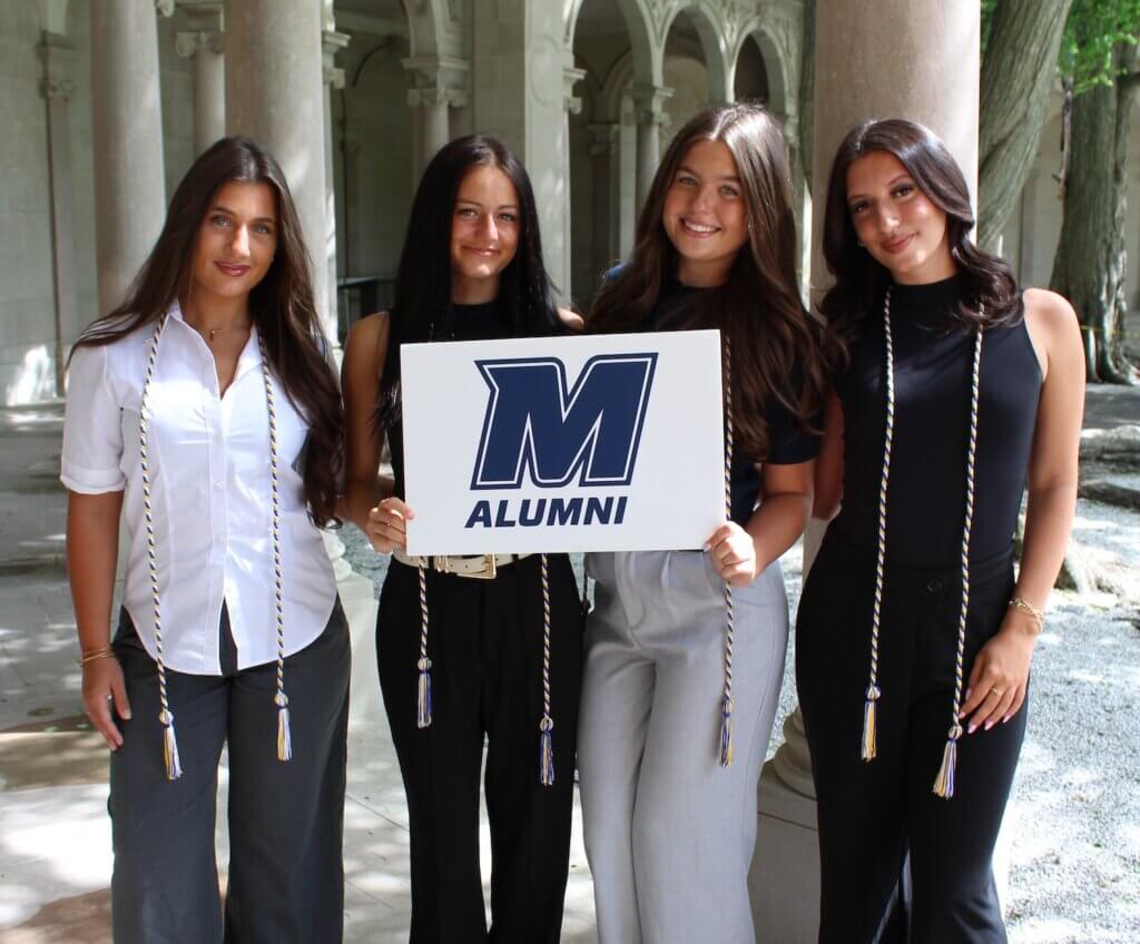 Four women holding up a Monmouth Alumni sign in front of stone columns in a garden.