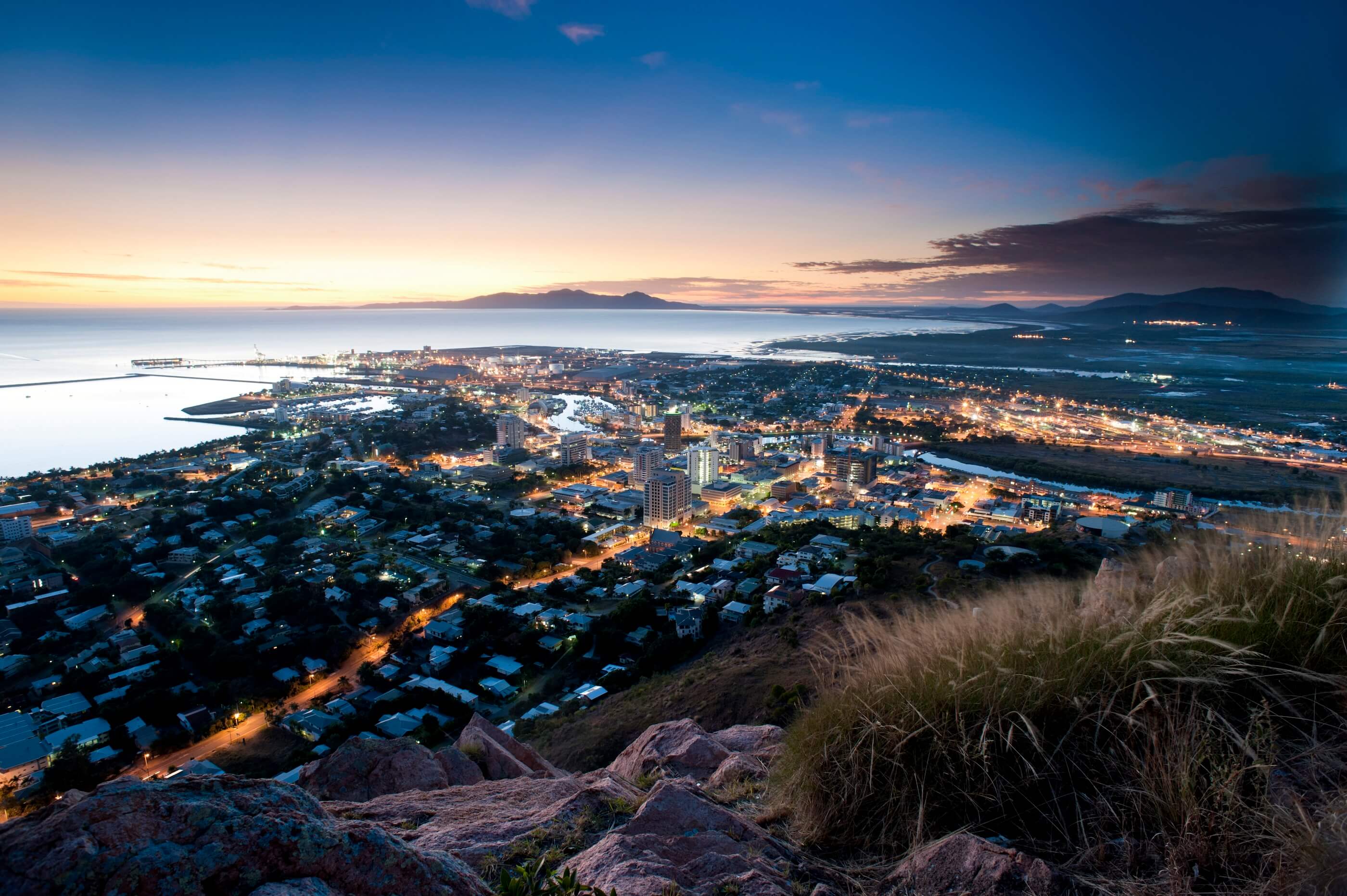 Nighttime shot of coastal city, small and tall buildings mixed together, lights illuminating roads. Sun rests on the horizon.
