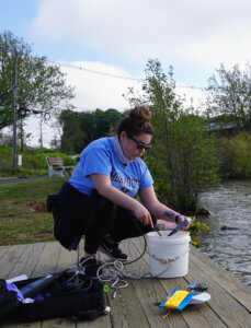 A student bending over a sample buck near a body of water. 