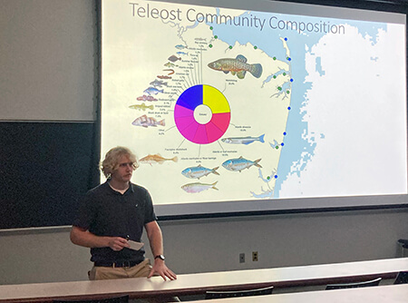 A presenter standing before a slide showing a map and a pie chart.
