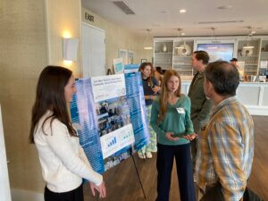 Two students speaking with a visitor while standing before a poster mounted on an easel. 