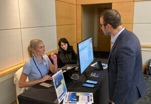 Two women seated at a table demonstrating software on a computer monitor to a visitor.