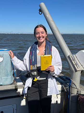 A woman standing aboard a vessel holding a book titled "All-Weather Environmental Field Book"