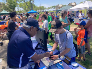 A man standing at a table teaching a boy how to use software on a phone at a crowded outdoor event.