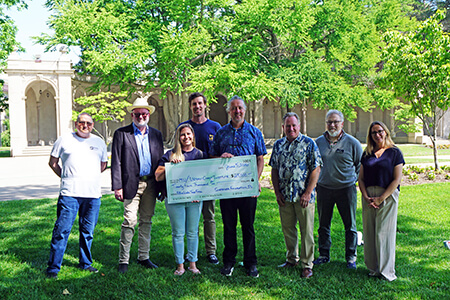 Eight men an women standing outside on a grassy garden area with a large check.