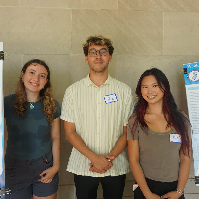 Three students standing together for a group photo between two research posters on easels.