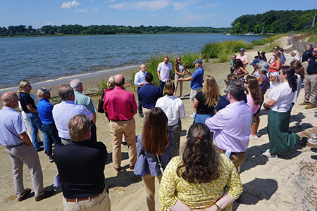 A crowd of a few dozen people listening to a speaker on a bay beach.