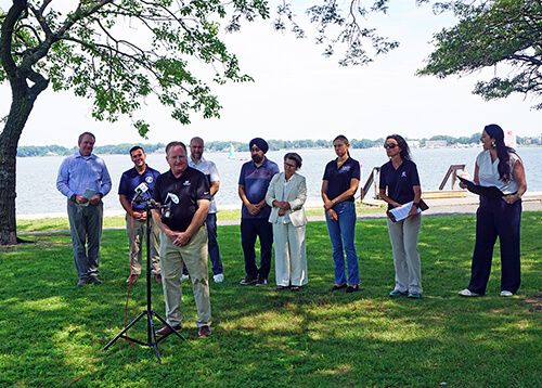 A man speaking at a microphone with eight people standing behind him at a waterfront.