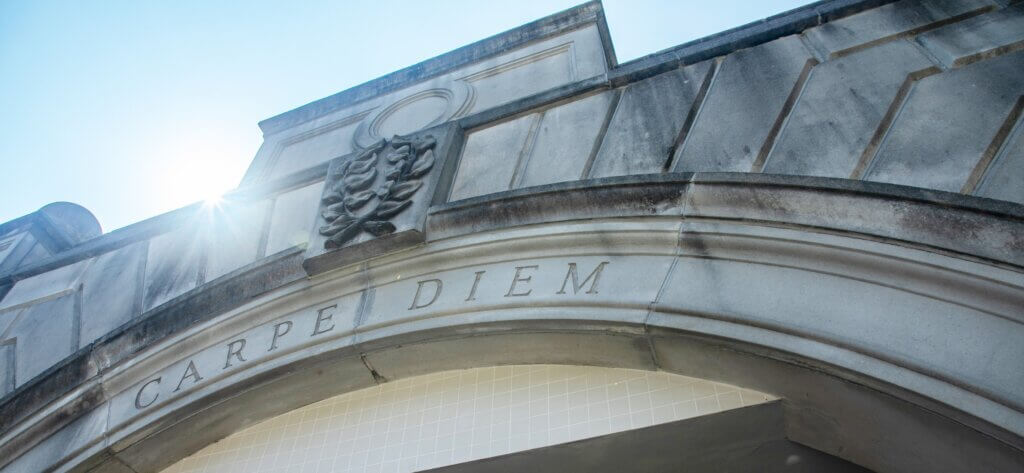The words Carpie Diem are carved into a stone arch with the sun behind it against a blue sky